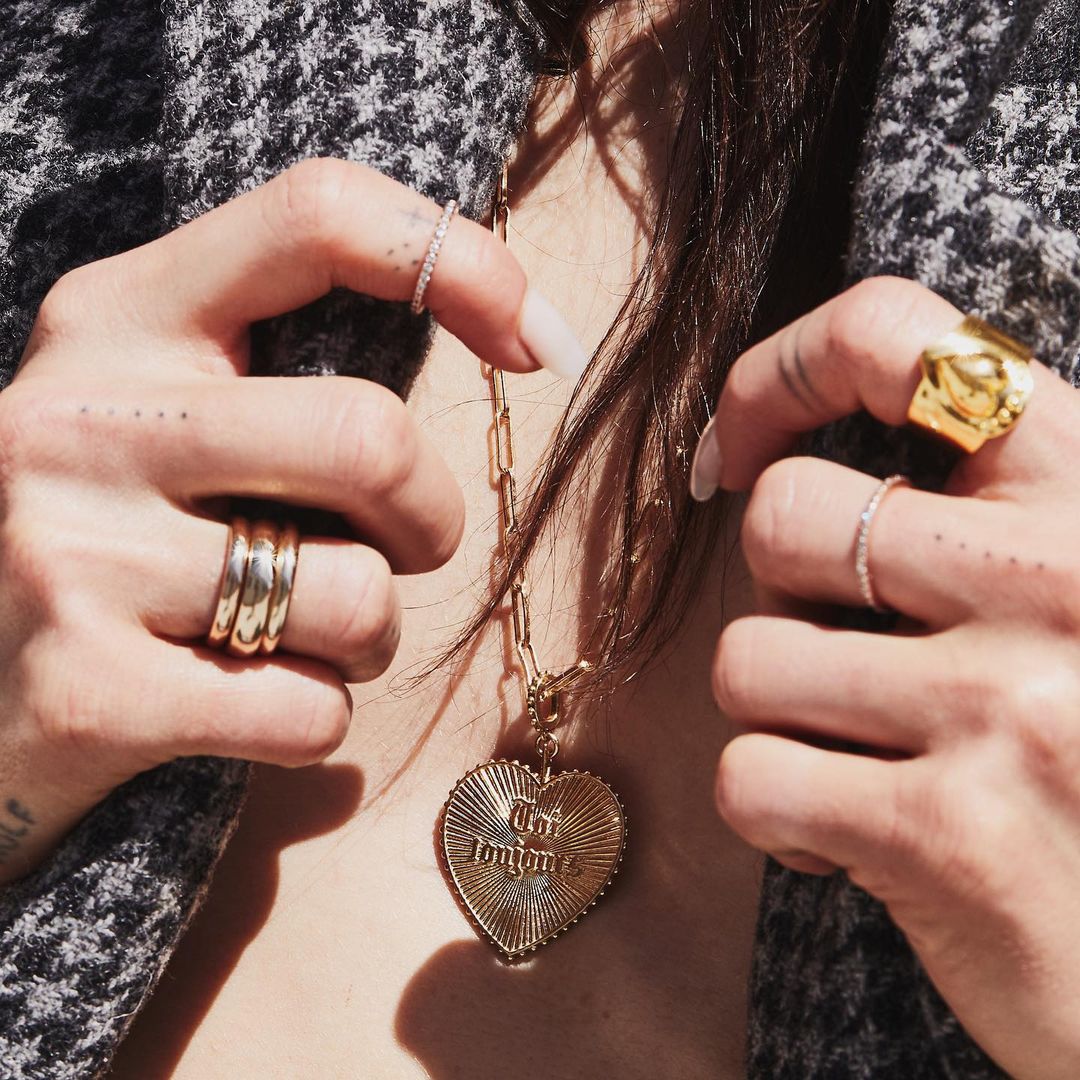 Girl holding her jacket open revealing her Toi Toujours Charm Heart Necklace with Puff Heart Cigar Band and Gold Stacked Bands